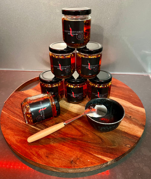 Six jars of fiery red chili oil are arranged in a pyramid and on a round wooden serving board, with a small black bowl and a wooden-handled spoon. The jars have black lids and black labels with white and red text.